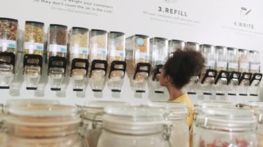 Young African American woman is choosing and shopping for organic products in refill store with reusable bag, zero-waste grocery, and plastic-free, eco environment-friendly, sustainable lifestyles.