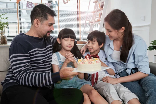Happy Asian Thai family, young daughter is surprised with birthday cake, blows candle, pray and cheerful celebrates party with parents together in living room, wellbeing domestic home event lifestyle.