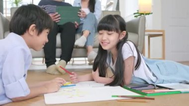 Asian Thai siblings are lying on living room floor, drawing homework with colored pencils together, parents leisurely relax on a sofa, lovely happy weekend activity, and domestic wellbeing lifestyle.
