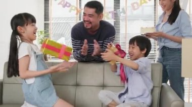 Happy Asian Thai family, young kids surprised by birthday cake, gift, blow out candle, and celebrate party with parents and siblings together in living room, wellbeing domestic home event lifestyle.