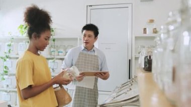 Asian male shopkeeper describes natural organic products to woman customer in refill store, zero-waste and plastic-free grocery, eco environment-friendly, sustainable lifestyles with a reusable shop.