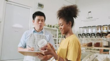 Asian male shopkeeper describes natural organic products to woman customer in refill store, zero-waste and plastic-free grocery, eco environment-friendly, sustainable lifestyles with a reusable shop.