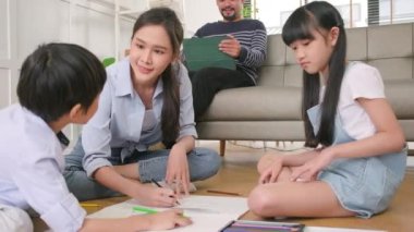 Asian Thai siblings and mum are sitting on living room floor, drawing with colored pencils together, dad leisurely relaxing on a sofa, lovely happy weekend activity, and domestic wellbeing lifestyle.