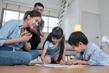 Asian Thai siblings and mum are sitting on living room floor, drawing with colored pencils together, dad leisurely relaxing on a sofa, lovely happy weekend activity, and domestic wellbeing lifestyle.