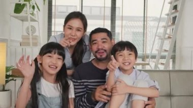 Happy Asian Thai parents, and children looking and talking to a camera, online video call with family via internet on sofa in home living room, lovely weekend, and domestic wellbeing lifestyle.