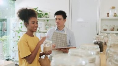 Asian male shopkeeper describes natural organic products to woman customer in refill store, zero-waste and plastic-free grocery, eco environment-friendly, sustainable lifestyles with a reusable shop.