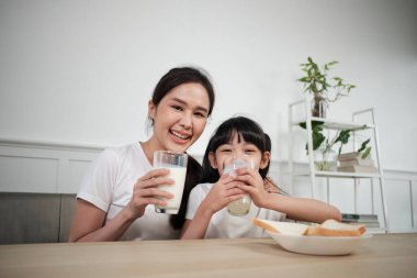 Sağlıklı Asyalı Tayland ailesinin portresi, mutlu kız ve genç anne kameraya bakıyor, yemek masasında birlikte taze süt ve ekmek içiyor, sağlıklı beslenme kahvaltısı yaşam tarzı.