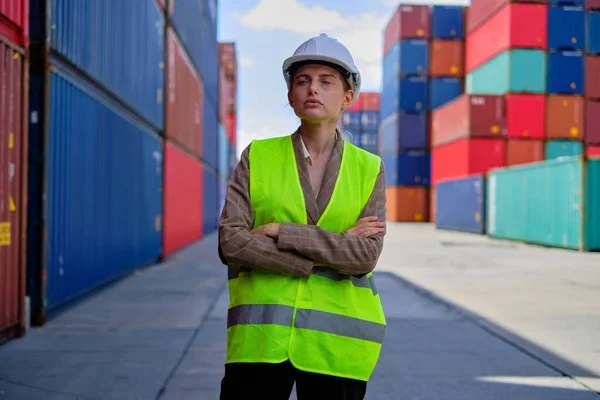 Portrait of professional White female worker in safety uniform and hardhat arms crossed and looks at camera works at logistics containers, import, and export cargo shipping, transportation industry.