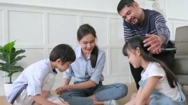 Asian Thai siblings and mum are sitting on living room floor, drawing with colored pencils together, dad leisurely relaxing on a sofa, lovely happy weekend activity, and domestic wellbeing lifestyle.
