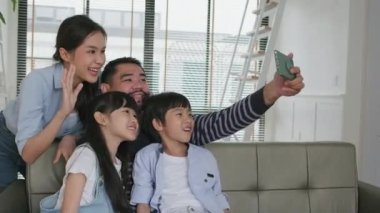 Happy Asian Thai parents, and children online video call with family via internet on sofa in home living room, lovely weekend, and domestic wellbeing lifestyle with internet technology mobile phone.