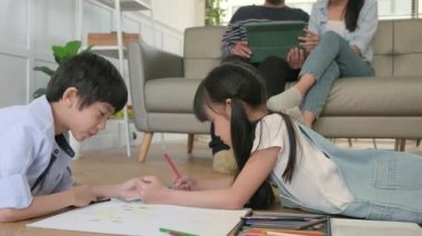 Asian Thai siblings are lying on living room floor, drawing homework with colored pencils together, parents leisurely relax on a sofa, lovely happy weekend activity, and domestic wellbeing lifestyle.