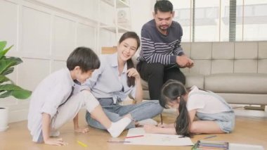 Asian Thai siblings and mum are sitting on living room floor, drawing with colored pencils together, dad leisurely relaxing on a sofa, lovely happy weekend activity, and domestic wellbeing lifestyle.