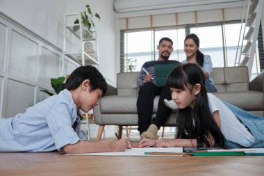 Asian Thai siblings are lying on living room floor, drawing homework with colored pencils together, parents leisurely relax on a sofa, lovely happy weekend activity, and domestic wellbeing lifestyle.