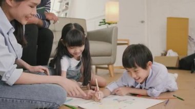 Asian Thai siblings and mum are sitting on living room floor, drawing with colored pencils together, dad leisurely relaxing on a sofa, lovely happy weekend activity, and domestic wellbeing lifestyle.