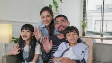 Happy Asian Thai parents, and children looking and talking to a camera, online video call with family via internet on sofa in home living room, lovely weekend, and domestic wellbeing lifestyle.