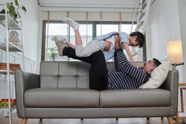 Asya Tayland ailesi birlikte, baba uzanıp oğluyla oynayarak eğleniyor. Oturma odasındaki kanepede uçan uçak gibi kollarını açıyor, güzel boş zamanları, güzel bir hafta sonu, ev hayatı gibi..