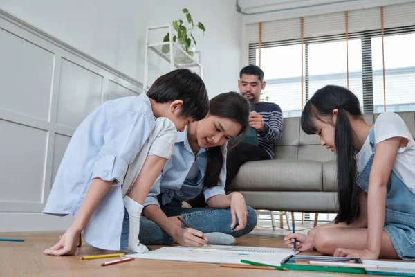 Asian Thai siblings and mum are sitting on living room floor, drawing with colored pencils together, dad leisurely relaxing on a sofa, lovely happy weekend activity, and domestic wellbeing lifestyle.