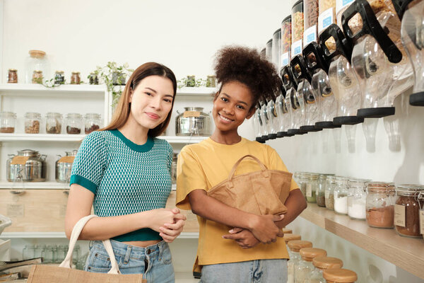 Two female customers look at camera and smile, shopping for organic products in refill store with reusable bags, zero-waste grocery, and plastic-free, environment-friendly, sustainable lifestyles.