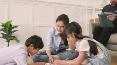 Asian Thai siblings and mum are sitting on living room floor, drawing with colored pencils together, dad leisurely relaxing on a sofa, lovely happy weekend activity, and domestic wellbeing lifestyle.