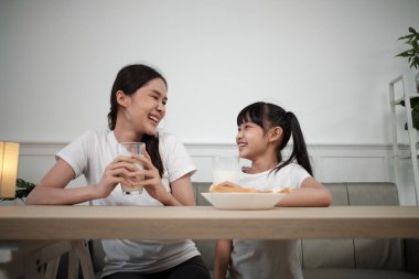 A Healthy Asian Thai family, little daughter, and young mother drink fresh white milk in glass and bread joy together at a dining table in morning, wellness nutrition home breakfast meal lifestyle.
