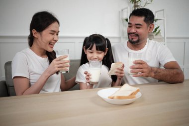 Sağlıklı bir Asyalı Taylandlı aile, küçük bir kız çocuğu ve genç ebeveynler sabah birlikte yemek masasında taze beyaz süt ve ekmek sevinci içerler, sağlıklı beslenme kahvaltısı ev yemeği yaşam tarzı..