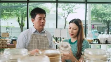 Asian male shopkeeper describes natural organic products to woman customer in refill store, zero-waste and plastic-free grocery, eco environment-friendly, sustainable lifestyles with a reusable shop.