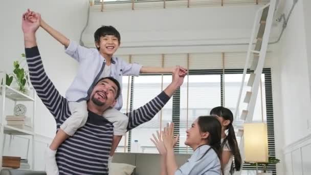 アジア系タイ人の家族と一緒に 家庭のリビングルームで肩の上に息子を運んだり抱いたりすることで お父さんは子供やお母さんと遊びやお茶をしたり 楽しい週末を過ごすことができます — ストック動画