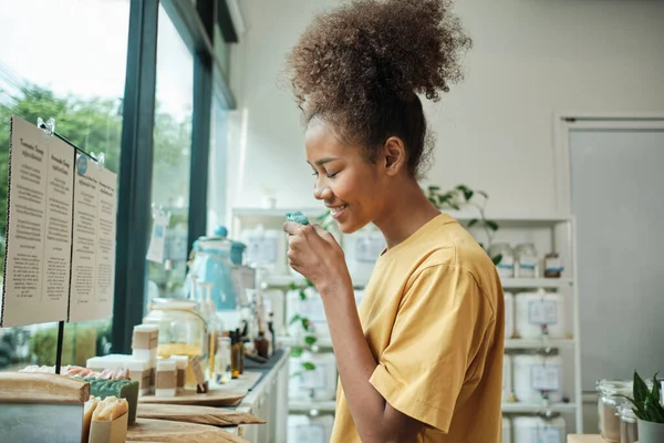 Young African American woman smells organic soaps, chooses and shops for reusable appliance products in refill store, zero-waste grocery, and plastic-free, environment-friendly, sustainable lifestyle.