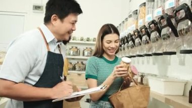 Asian male shopkeeper describes natural organic products to woman customer in refill store, zero-waste and plastic-free grocery, eco environment-friendly, sustainable lifestyles with a reusable shop.