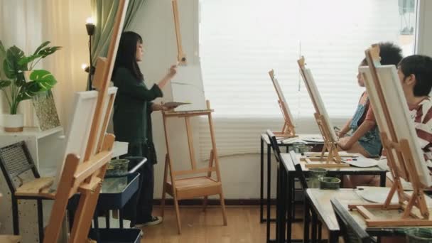 アジアの女性教師が 小学校のスタジオ教育でスキルを使って創造的に学ぶ 教室でのキャンバス上のアクリルカラー絵具について子供たちに教え 実証しています — ストック動画