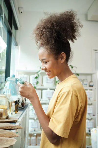 A young African American woman chooses and shops for reusable and recycled appliance products, soap in refill store, zero-waste grocery, and plastic-free, environment-friendly, sustainable lifestyle.