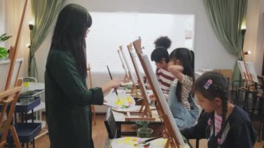 Kadın bir Asyalı ile öğrenen bir grup çok ırklı çocuk sanat sınıfındaki tuvale akrilik renkli resim çizmeyi öğretiyor, ilkokul stüdyo eğitiminde yaratıcı bir şekilde öğreniyorlar..