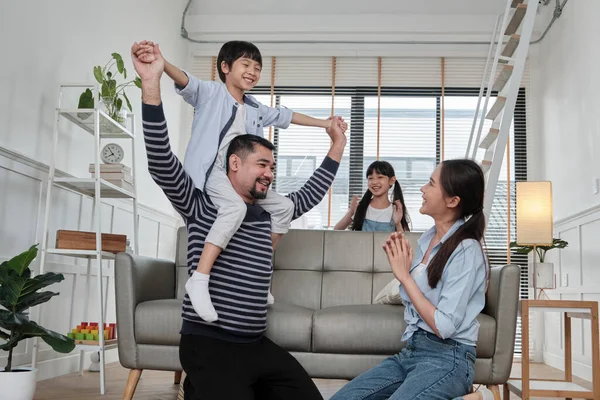 Asian Thai family together, dad plays and teases with children and mum by carrying and holding son on shoulders in home living room, happy leisure times, lovely weekend, wellbeing domestic lifestyle.