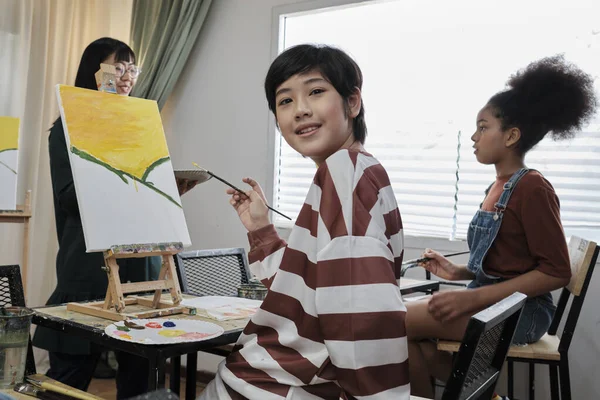 Portrait of Asian boy looking at camera and smiling with acrylic color picture painted on canvas in art classroom and creatively learning with talents and skill at elementary school studio education.