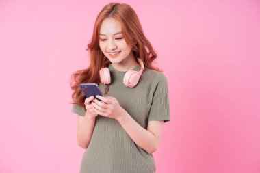 Genç Asyalı kadın akıllı telefon kullanıyor ve pembe arka planda müzik dinliyor.