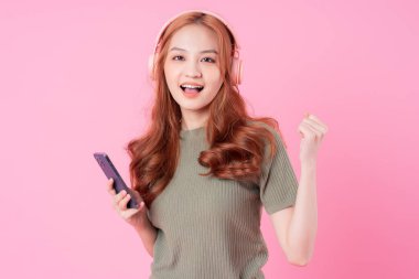 Genç Asyalı kadın akıllı telefon kullanıyor ve pembe arka planda müzik dinliyor.