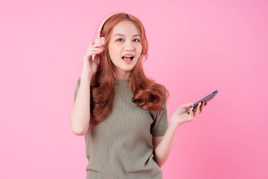 Genç Asyalı kadın akıllı telefon kullanıyor ve pembe arka planda müzik dinliyor.