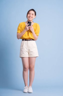 Mavi arka planda akıllı telefon kullanan genç Asyalı kadının tam boy görüntüsü