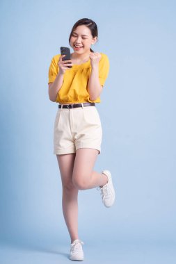 Mavi arka planda akıllı telefon kullanan genç Asyalı kadının tam boy görüntüsü