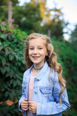 Neşeli, sarı saçlı, mavi gözlü bir kızın portresi yemyeşil çalıların arka planında. Kız mavi bir kot ceket giyiyor.