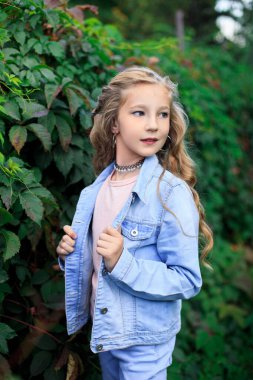 Neşeli, sarı saçlı, mavi gözlü bir kızın portresi yemyeşil çalıların arka planında. Kız mavi bir kot ceket giyiyor.