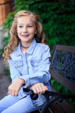 Parktaki bankta oturan neşeli, sarı saçlı, mavi gözlü bir kızın portresi. Kız mavi bir kot ceket giyiyor.