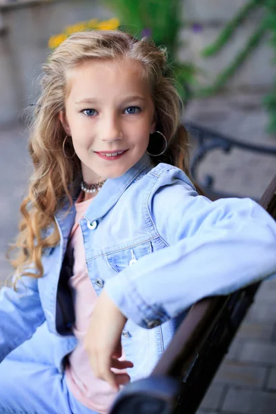 公園のベンチに座って楽しいブロンドの髪の青い目の少女の肖像画 女の子は青いデニムジャケットを着ている — ストック写真