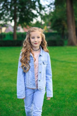 Neşeli sarı saçlı, mavi gözlü bir kızın portresi, yeşil çimlere karşı mavi kot ceketli bir kız.