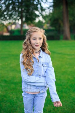 Neşeli sarı saçlı, mavi gözlü bir kızın portresi, yeşil çimlere karşı mavi kot ceketli bir kız.