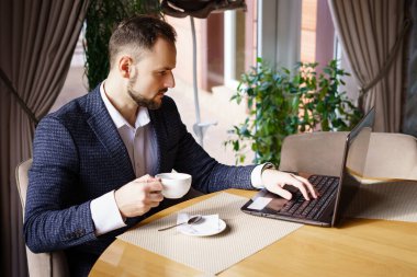 Takım elbiseli genç bir adam restoranda bir masada oturuyor. Bir restoranda ya da kafede bilgisayarında çalışırken kahve içen bir iş adamı.