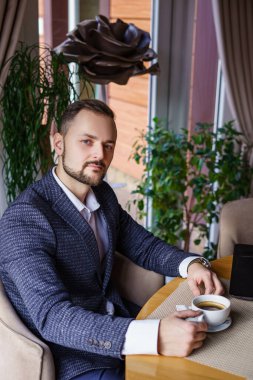A young man with a beautiful beard is drinking coffee at a table in a restaurant. A man in a dark business suit in a cafe