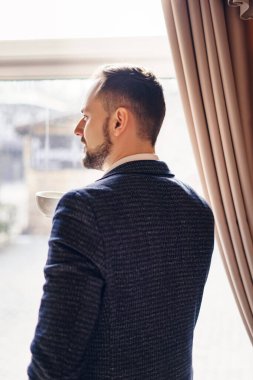 Takım elbiseli genç bir adam pencerenin yanında kahve içiyor. Kafede sakallı genç bir adam..