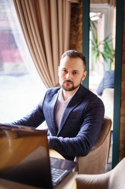 Takım elbiseli bir adam restorandaki bir masada dizüstü bilgisayarında oturuyor ve beyaz bardaktan kahve içiyor. Sakallı genç bir adam restoranda bir masada.. 