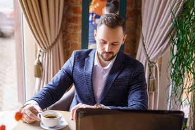 Takım elbiseli bir adam restorandaki bir masada dizüstü bilgisayarında oturuyor ve beyaz bardaktan kahve içiyor. Sakallı genç bir adam restoranda bir masada.. 